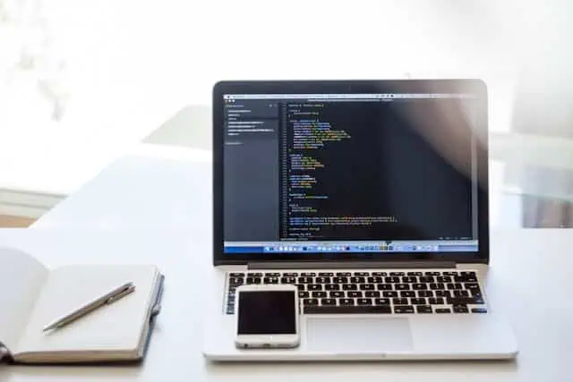 A laptop showing code on the screen, a smartphone and a notebook with a pen on a desk.