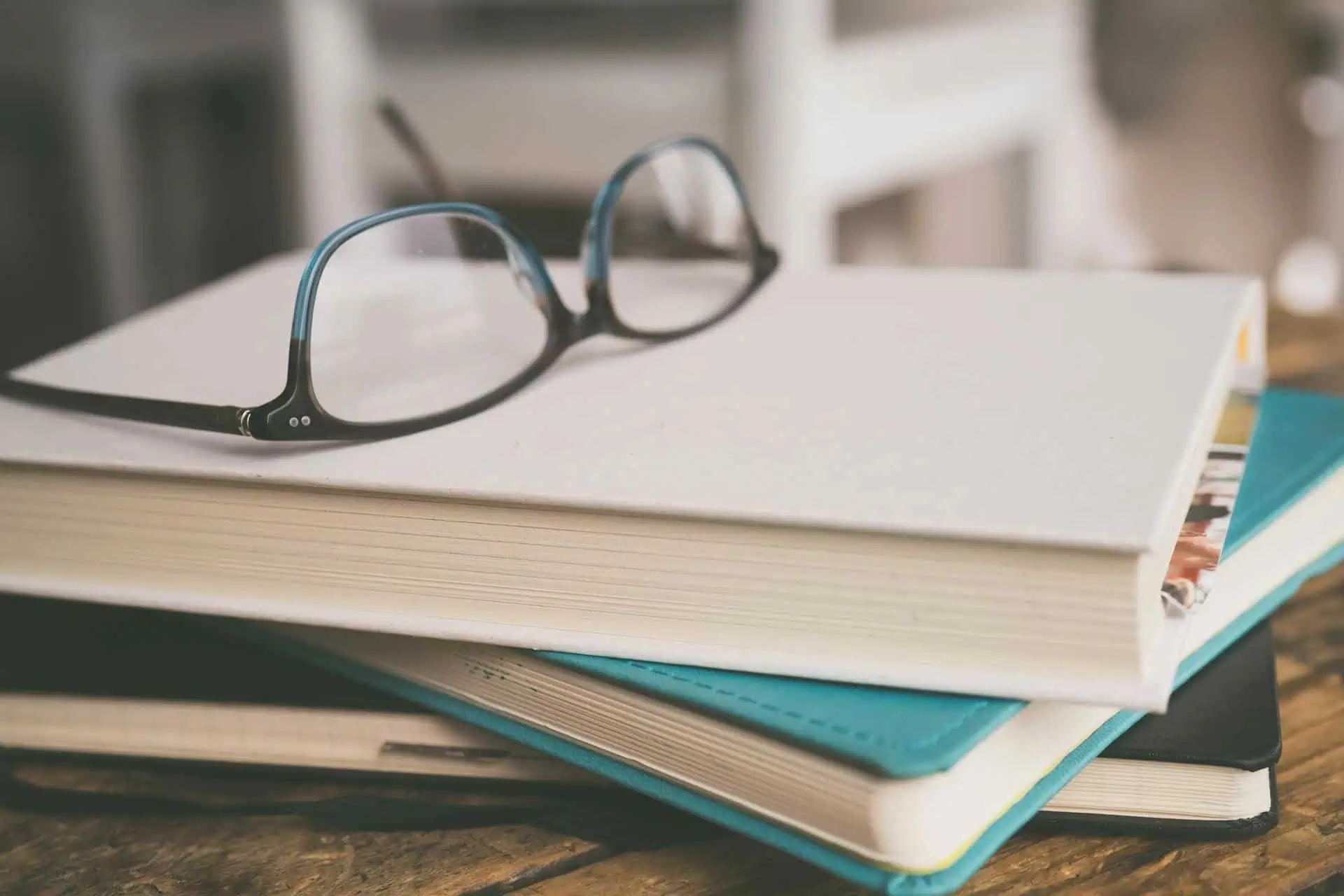 Three stacked books with a pair of glasses on top, on a wooden table or desk.